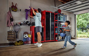 A person organizes sports equipment in a garage using red and black CRAFTSMAN storage cabinets and shelves. A child holding a helmet approaches. Various items like balls, rackets, and bags are visible, and the garage door is open to a green yard.